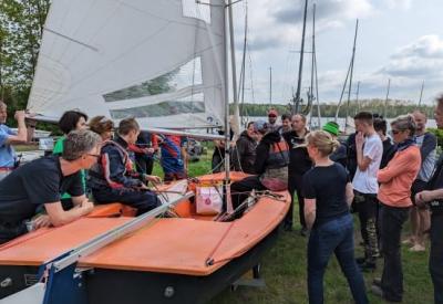 Eine orangene Ixylonjolle an Land mit vielen Zuschauern drum herum